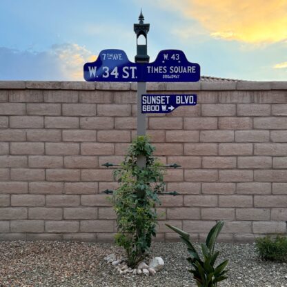 A photograph of two New York humpback street signs and one Los Angeles shotgun street sign mounted to a post in the yard of a desert home.